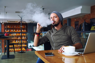Young handsome hipster man with beard sitting in cafe with a cup of coffee, vaping and releases a...