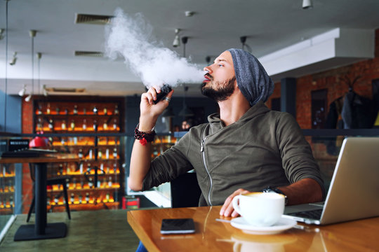 Young handsome hipster man with beard sitting in cafe with a cup of coffee, vaping and releases a cloud of vapor. Working at laptop and having a little break. With copy space.