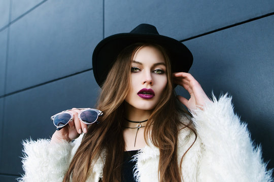Outdoor closeup portrait of young fashionable woman posing on street, on blue background. Model with beautiful makeup, looking at camera, holding sunglasses, wearing stylish hat, white fake fur coat