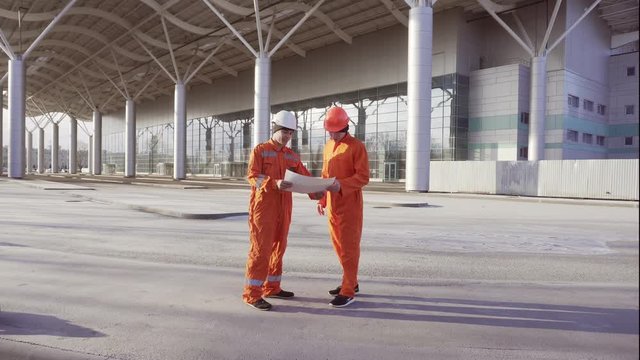Two construction workers in orange uniform and hardhats meeting each other at the bulding object and examining the constructed building together. Teamwork concept
