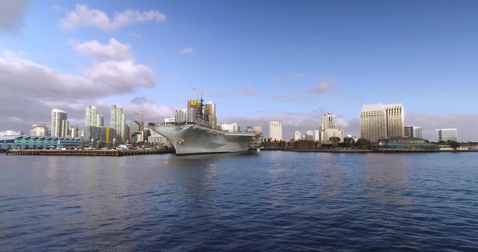 A wide, dramatic daytime establishing shot of the San Diego city skyline as seen from the bay. The USS Midway aircraft carrier is in the foreground. Part 2 of 2.	 	