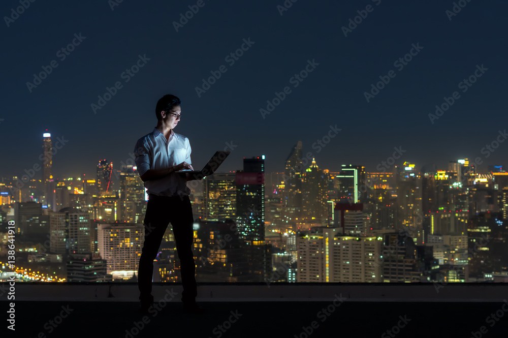 Wall mural asian businessman standing and using the laptop over the cityscape background at night time, busines