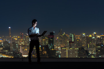 Asian businessman standing and using the laptop over the cityscape background at night time,...