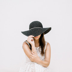 Young pretty woman in white dress with black hat near white wall.