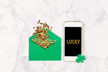 St. Patrick's Day concept.  Flat lay stylish set: gold confetti in the envelope, clover leaf and smartphone. Mockup template. View from above