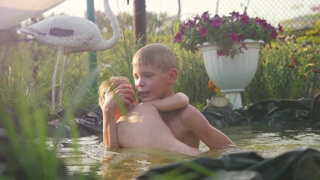Two happy brothers swimming in a pool in summer garden in slowmotion. Younger brother holds younger brother have a fun time. 1920x1080