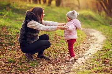 Fototapeta premium Young mother playing with daughter on autumn forest background. Family walking in park. Bright back light.