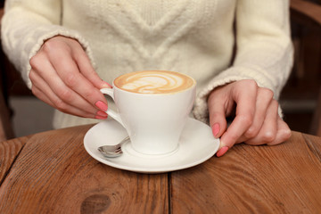 Close-up female hands holding cup with coffee cappuccino with foam with nice pattern. Perfect red gel polish manicure. Wood natural vintage table. Creative color warm post processing