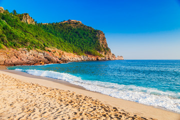 Naklejka premium Plaża Kleopatry na wybrzeżu morskim z zielonymi skałami na półwyspie Alanya, dystrykt Antalya, Turcja. Piękny słoneczny krajobraz dla turystyki z czystą wodą i piaskiem. Zamek Alanya na klifie