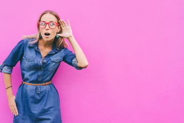 Gossip concept. Colorful portrait of amazed young woman with palm near her ear. Pink background.