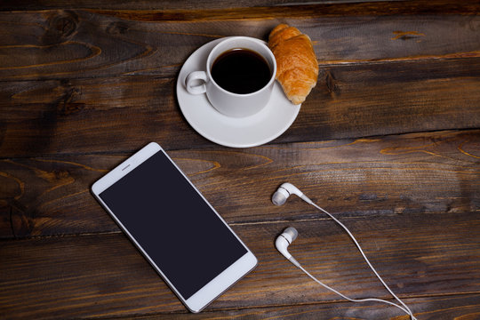 White mobile phone on wooden wooden background with headphones, cup of coffee and croissant