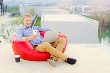 Freelance businessman. Young handsome man working on laptop and using smartphone while sitting on the red beanbag.