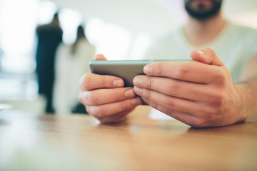 Side view of young bearded man,dressed incasual wear,sitting at round wooden table in cafe with modern interior and is holding smartphone.