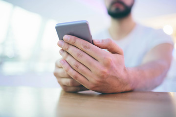 Side view of young bearded man,dressed incasual wear,sitting at round wooden table in cafe with modern interior and is holding smartphone.