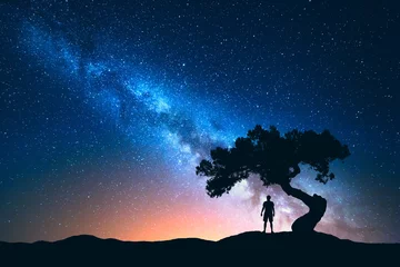 Poster Night landscape with Milky Way and silhouette of man under the tree growing from the rock on the mountain. Nature background with starry sky and beautiful galaxy. Blue Milky Way and man. Universe © den-belitsky