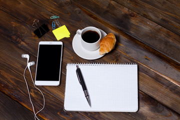 White mobile phone on wooden wooden background with headphones, cup of coffee, croissant and Stationery appliances