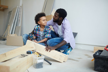 Cute boy talking to father while drilling wooden plank during house renovation