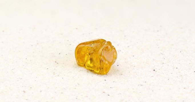 Amber gem rotating on white sand surface. Gemstone formed through fossilization of tree resin with small mosquitos inside. Clip contains seamless loop.
