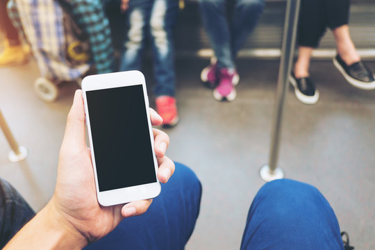 Mockup image of hand holding white mobile phone with blank black screen in subway with many people in background