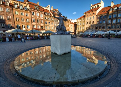 Statue of Mermaid (Syrenka - symbol of Warsaw) at the Old Town Market Square against tenements and restaurants in warm lights of sunset in Warsaw, Poland