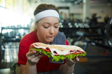 Portrait of cute overweight woman licking her lips wanting to eat huge fattening sandwich while working out in gym, struggling to stay in shape