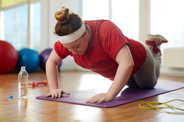 Portrait of young obese woman working out on yoga mat in sunlit fitness studio: performing knee push up exercise with effort to lose weight