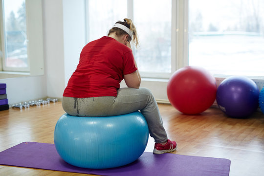 Back View Portrait Of Sweaty Overweight Woman Working Out In Fitness Studio: Sitting On Big Fitness Hanging Her Head Looking Upset And Exhausted