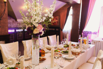 Tall glass vase with white and violet flowers stands in the middle of reach served dinner table