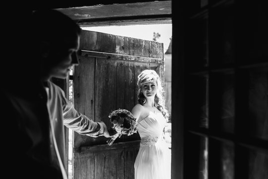 Groom reaches out his hand to bride standing before wooden door in rays of sun