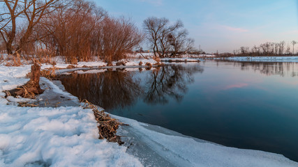 evening winter landscape. February thaw. Coast of the river with the ice edge and the vegetation under the snow