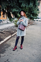 Young model girl in a gray coat and black hat with leather handbag on shoulders posed on street of city.