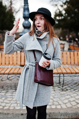 Young model girl in a gray coat and black hat with leather handbag on shoulders stay with plastic cup of coffee at street of city.