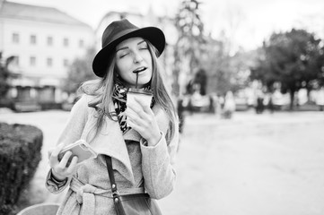 Young model girl in a gray coat and black hat with leather handbag on shoulders stay with plastic cup of coffee and making selfie at street of city. Black and white photo.