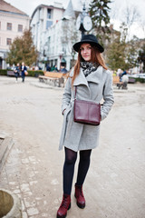 Young model tourist girl in a gray coat and black hat with leather handbag on shoulders posed at street of city.