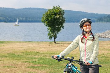 Female tourist cyclist  stands on shore of lake on  background of  mountains and enjoys a good day.