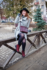 Young model girl in gray coat and black hat with leather handbag on shoulders posed against wooden beams at street of city.