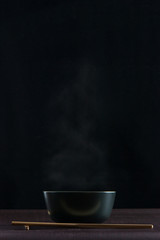 Black bowl with smoke boiling up and wooden chopsticks on wooden table with black background