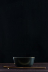 Black bowl with wooden chopsticks on wooden table with black background