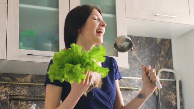 Slow motion of funny woman cook dance and sing with big ladle and lettuce while cooking in kitchen