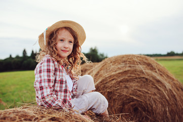 happy child girl in country style plaid shirt and hat relaxing on summer field with hay stacks