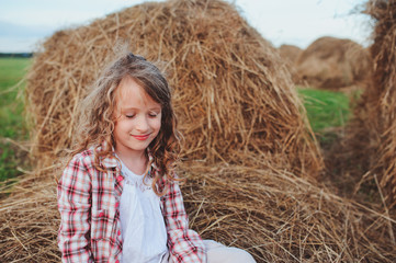 Naklejka na ściany i meble happy child girl in country style plaid shirt and hat relaxing on summer field with hay stacks