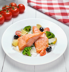 Baked salmon steak grilled with vegetables, olives, lemon and sauce on a white plate on a light wooden background.