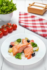 Baked salmon steak with vegetables, olives, lemon and sauce on a white plate on a light wooden background. Close-up.