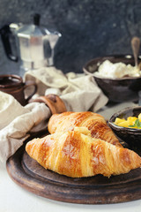 Breakfast with two croissant, butter, cup of coffee, cottage cheese and sliced mango fruit, served on serving wood board with textile napkin on white and gray concrete texture background. Close up.