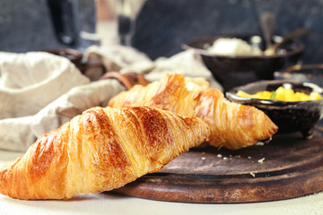 Breakfast with two croissant, butter, cup of coffee, cottage cheese and sliced mango fruit, served on serving wood board with textile napkin on white and gray concrete texture background. Close up.
