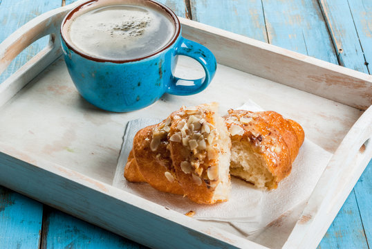 Summer breakfast: a large cup of hot coffee and a croissant with almonds. On the tray on a blue rustic table copy space