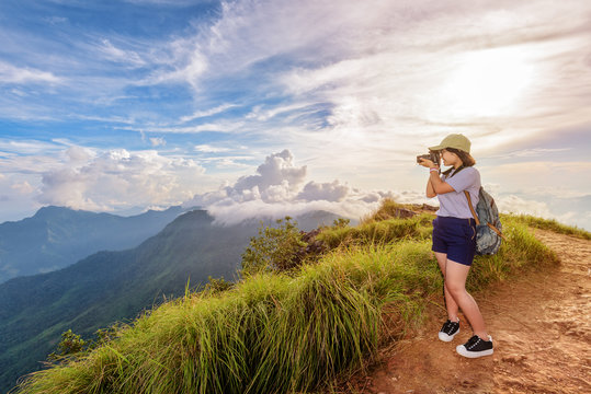 Hiker asian teen girl wear cap and eyeglass with happy holding dslr camera shooting photo beautiful nature in winter on peak mountain at sunset in Phu Chi Fa Forest Park, Chiang Rai, Thailand