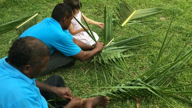 Fijian men teach young tourist girl how to create a basket from Coconut Palm