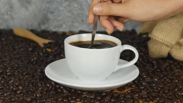4k of Close-Up Female Hand Stirring Cup Black Coffee - female hand using spoon to stir freshly brewed ground black coffee white cup saucer beside whole coffee beans, Coffee time