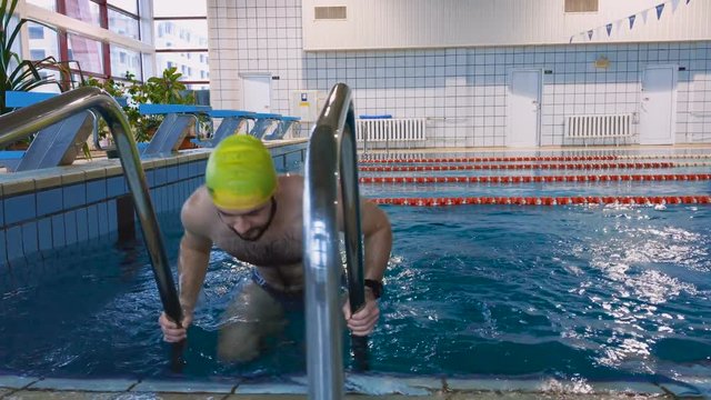 Muscular swimmer swims up the stairs and out of the pool. 4K.